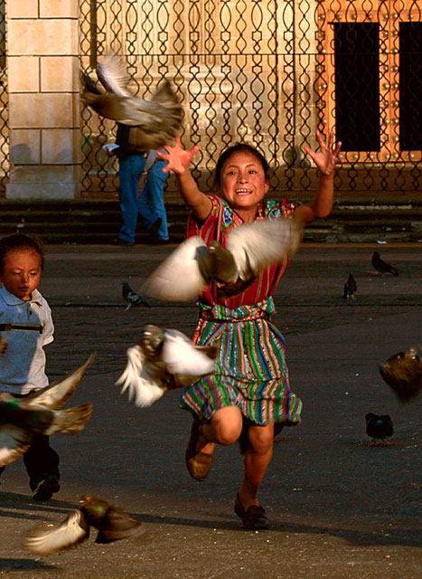 A Mayan child chases doves in front of the Cathedral pillars where the names of Mayans massacred during the war are memorialized, Guatemala City, Guatemala. Copyright © Donna De Cesare.Esta niña persigue palomas en frente de los pilares de la Catedral donde los nombres de los mayas masacrados durante la guerra están inscritos, Ciudad de Guatemala, Guatemala. Derechos reservados || Donna De Cesare.